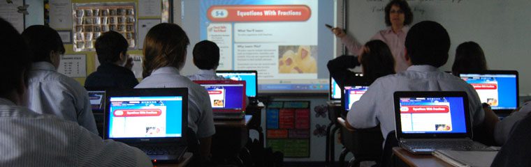 Students in a classroom on laptops facing a teacher presenting with a projector
